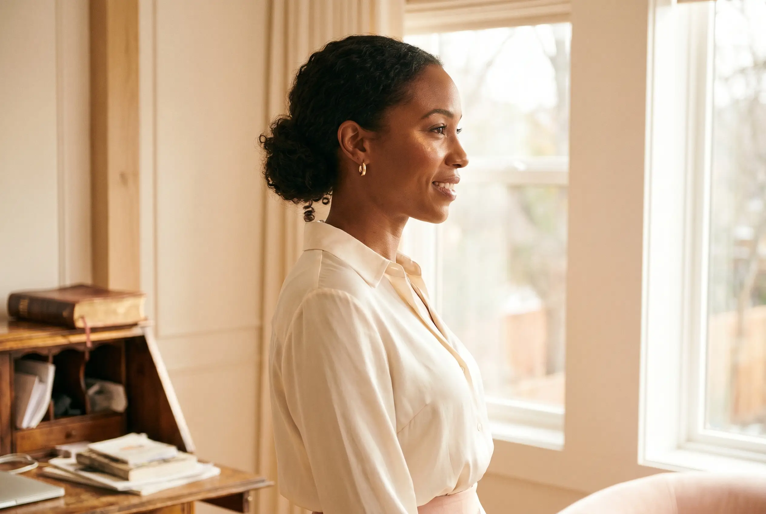 A Christian career woman in a cream silk blouse stands at her sunlit home-office window with a leather Bible on her desk in soft focus